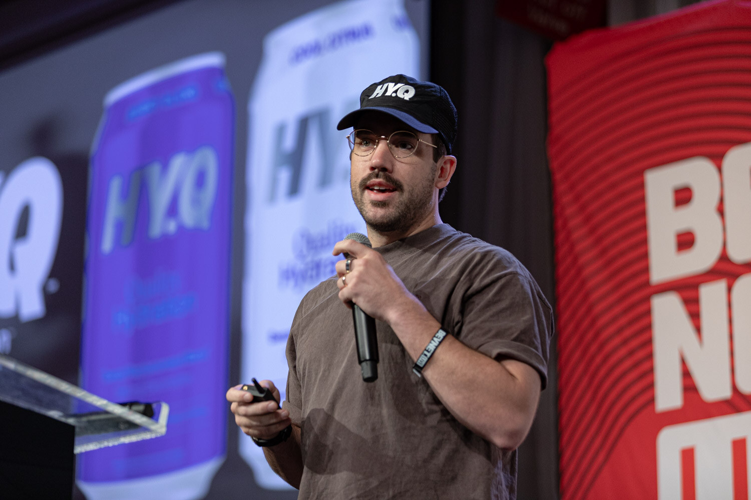 A man in a brown t-shirt and HY.Q cap holds a microphone and clicker in front of a screen showing HY.Q cans.