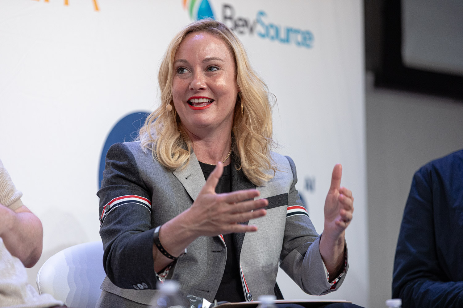 A woman with blonde hair in a gray blazer gestures with both hands while seated on stage.