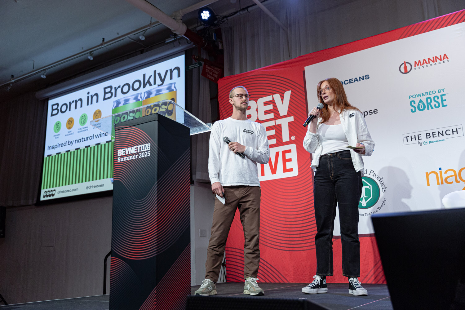 A man and a woman holding microphones stand next to a podium on a stage, with a slide reading Born in Brooklyn behind them.