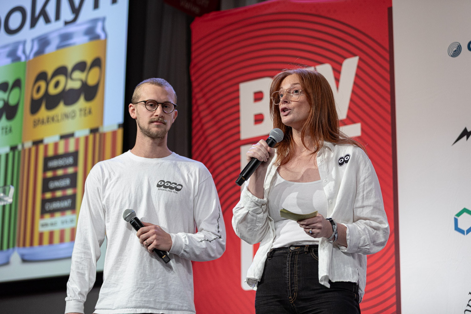 A man in a white long-sleeve shirt and a woman in a white jacket hold microphones in front of an OOSO slide.