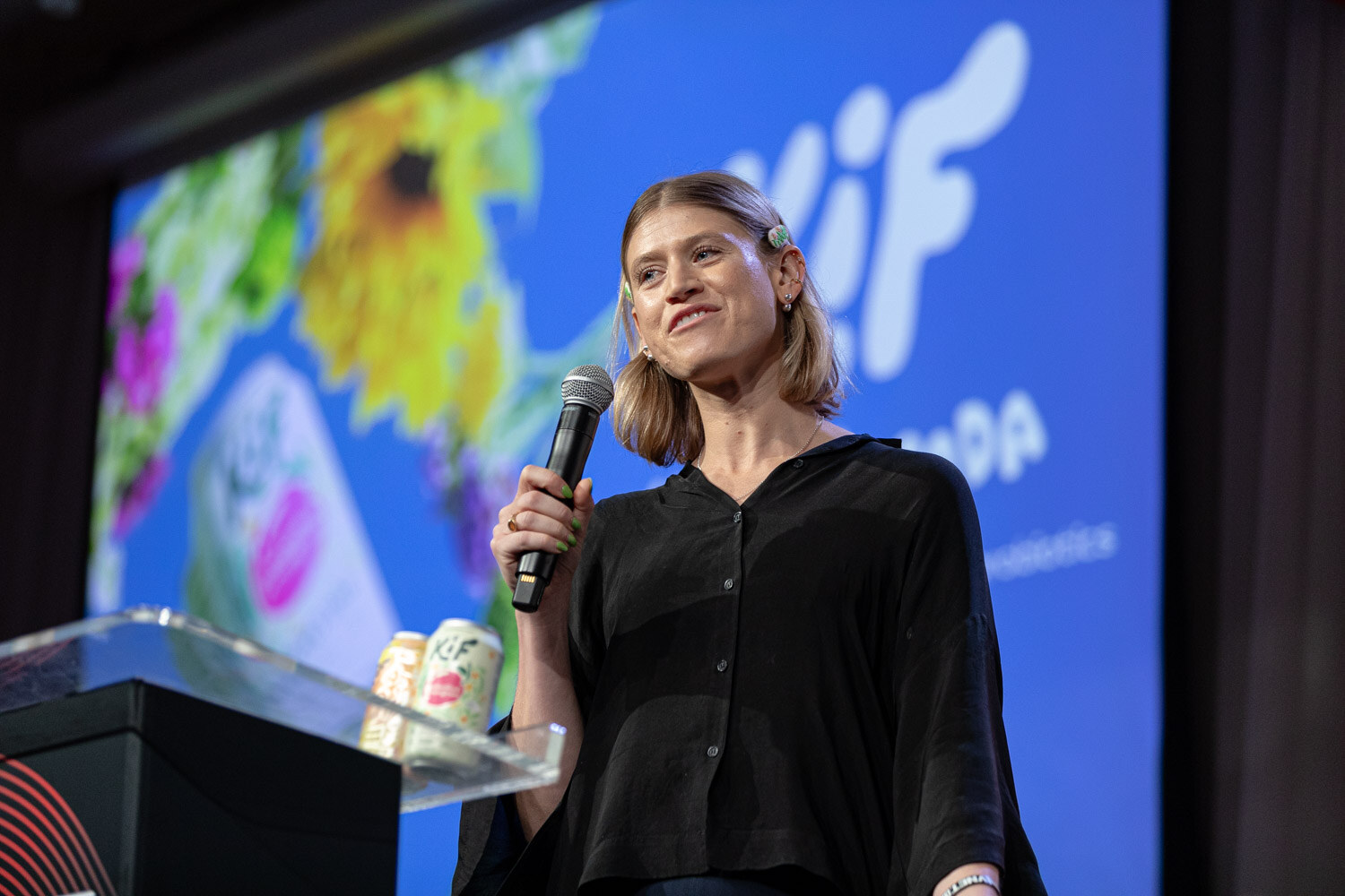 A woman in a black blouse holds a microphone near a podium with cans, in front of a bright Kif slide with flowers.