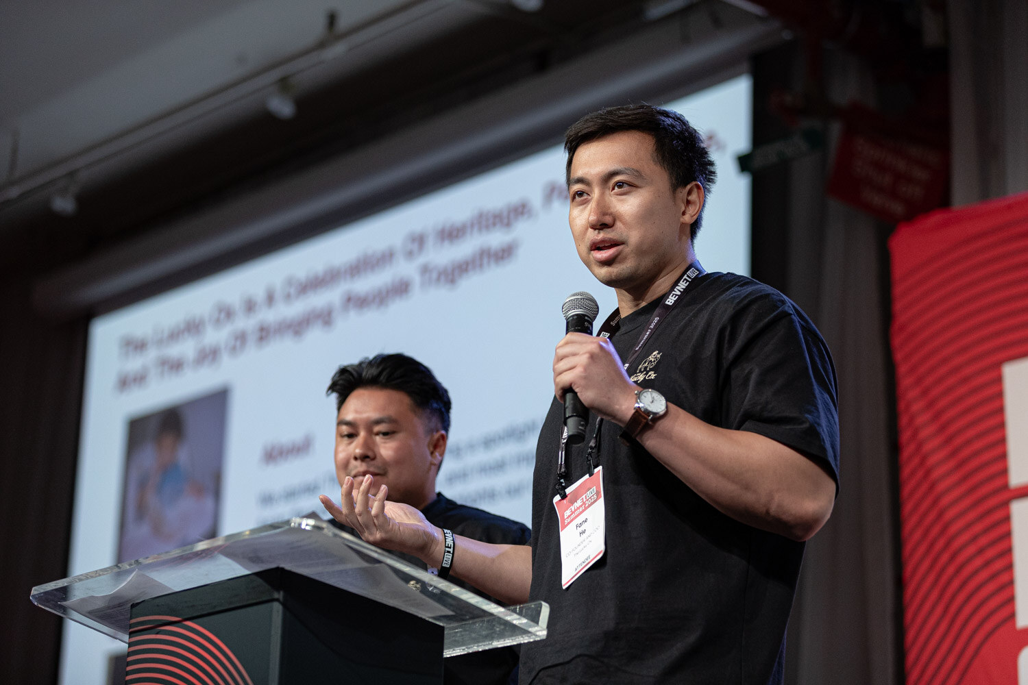 A man in a black t-shirt with a badge holds a microphone in front of a slide titled The Lucky Ox, with a second man behind him.