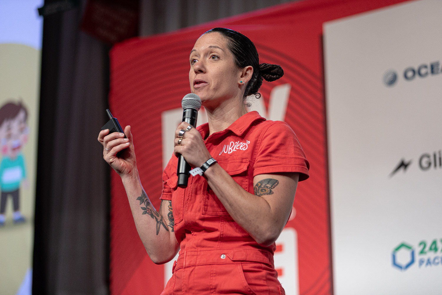A woman in a red jumpsuit with Jubilee's on the chest speaks into a microphone in front of a colorful slide.