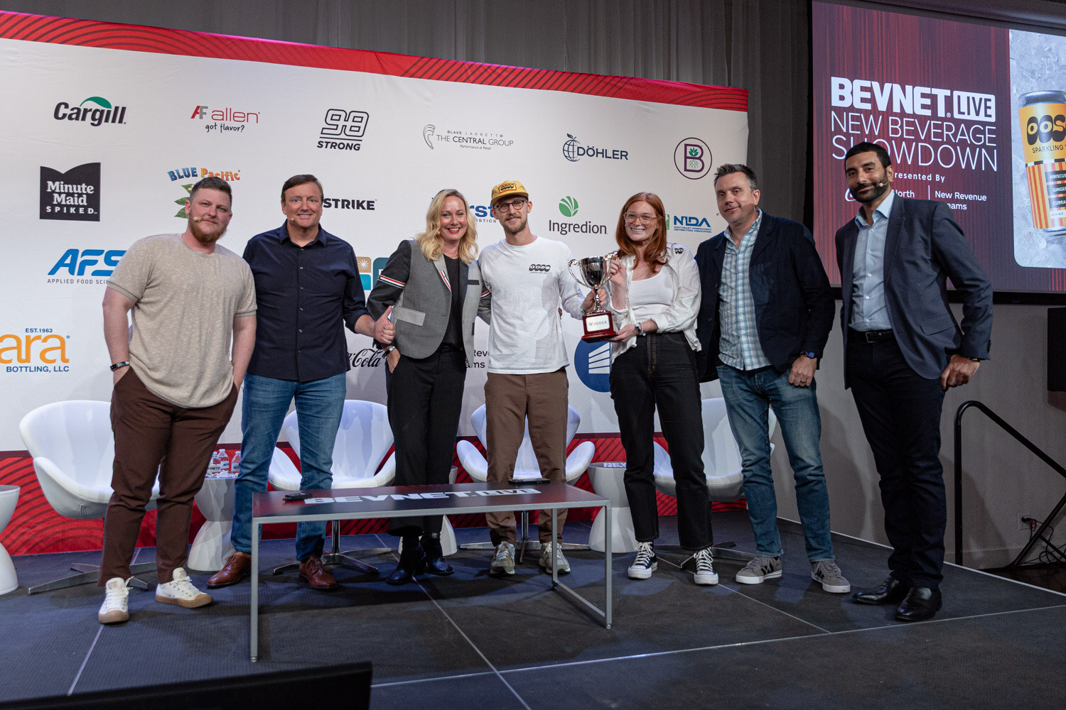 Seven people stand in a row on a stage holding a silver trophy in front of a New Beverage Showdown sign.