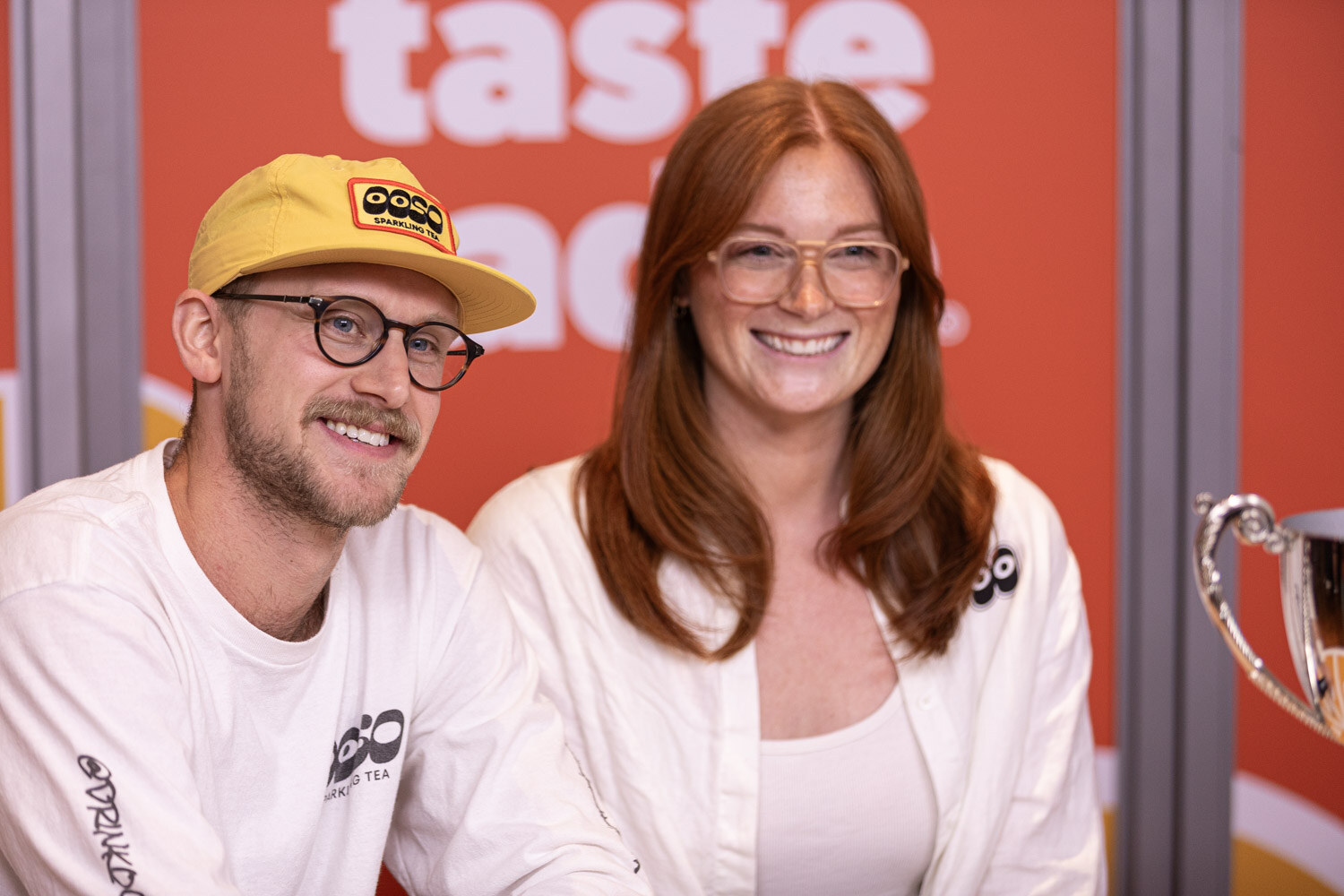 A man in a yellow cap with an OOSO logo and a woman with long red hair smile in front of an orange Taste Radio backdrop, with a silver trophy at the edge.