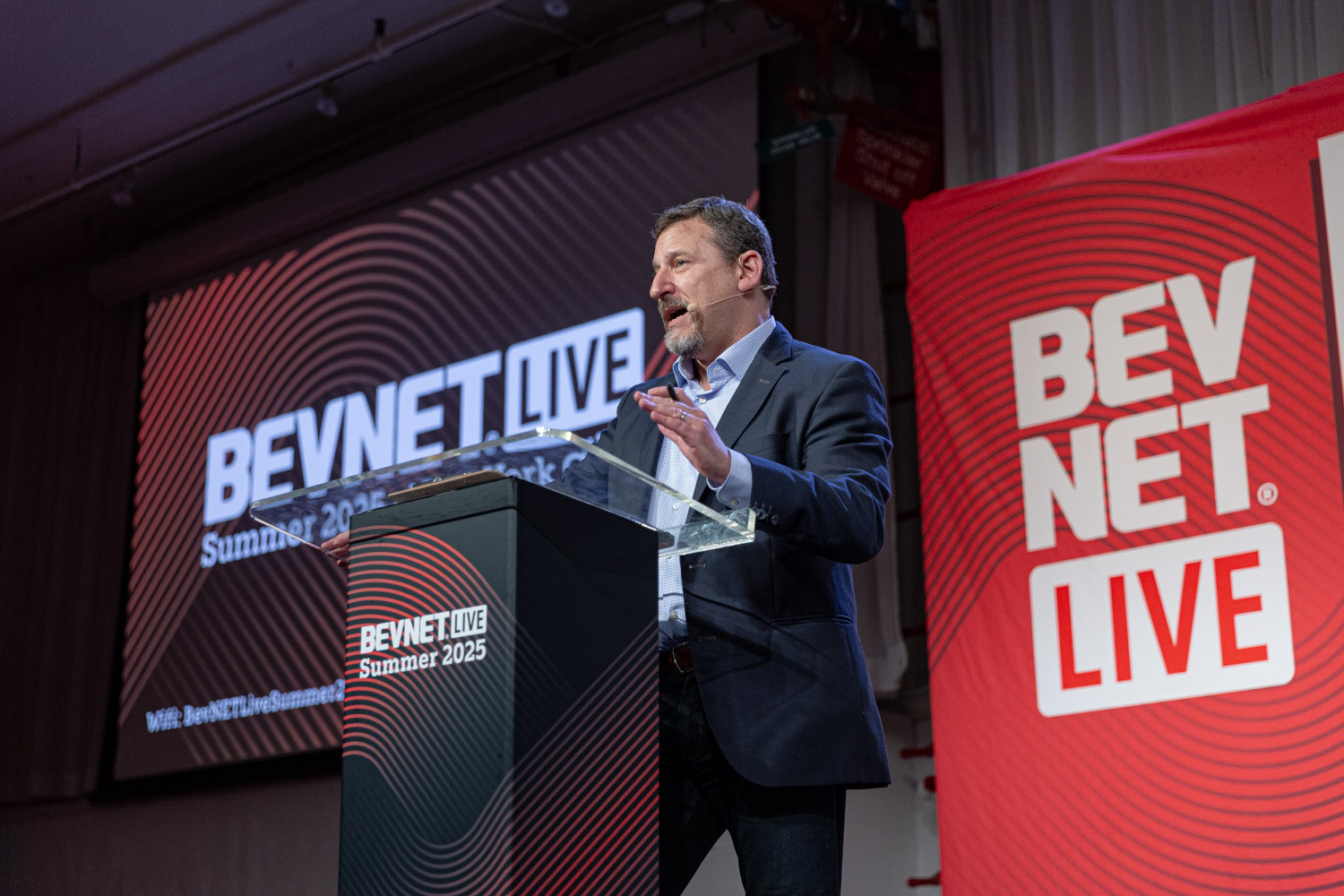 A man in a navy blazer stands at the podium next to a red BevNET Live banner and a stage screen.