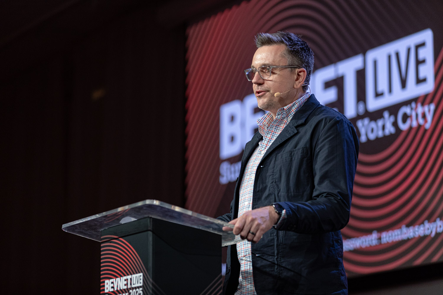 A man in a dark jacket and glasses speaks at a clear podium in front of a large BevNET Live New York City screen.