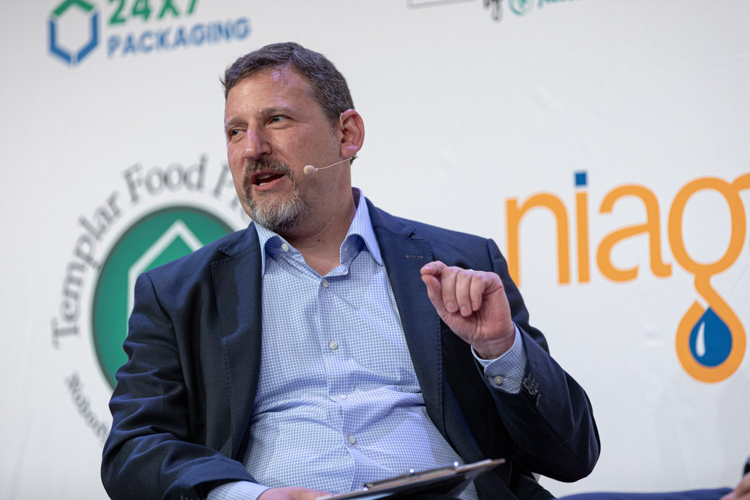 A bearded man in a navy blazer holds a clipboard and gestures while seated on stage in front of sponsor logos.
