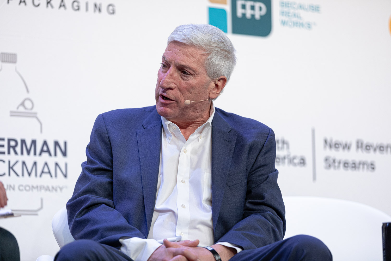 A gray-haired man in a blue blazer and open white shirt speaks while seated on a stage.