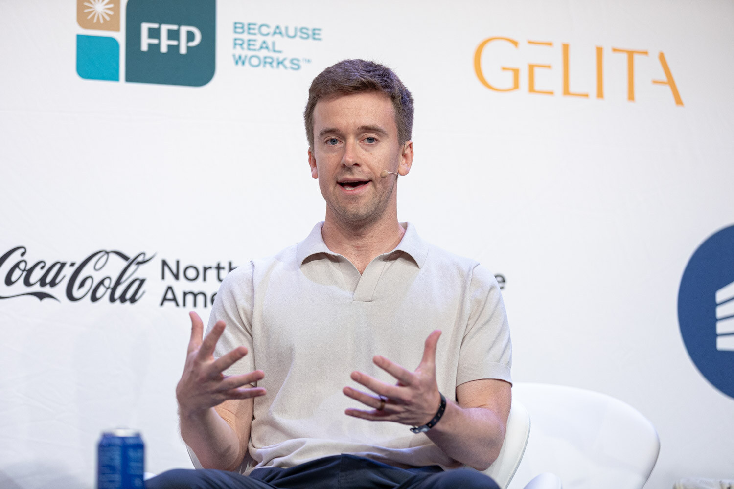 A young man in a cream polo speaks with both hands open on stage, with a Coca-Cola North America logo behind him.