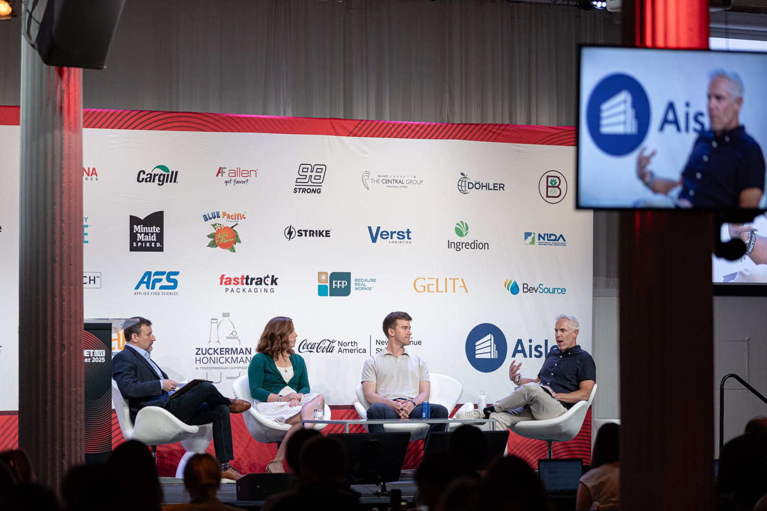 A wide shot of four people seated on a stage with a sponsor backdrop and a side screen showing one speaker.