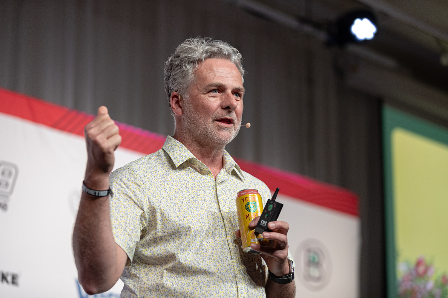 A gray-haired man in a patterned short-sleeve shirt holds a yellow can and clicker and raises a fist while speaking on stage.