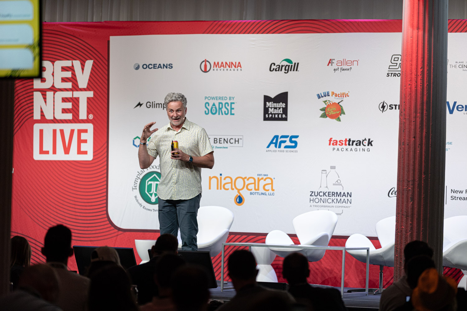 A wide shot of a man standing on a stage in front of a red BevNET Live banner and a wall of sponsor logos, with audience heads in the foreground.