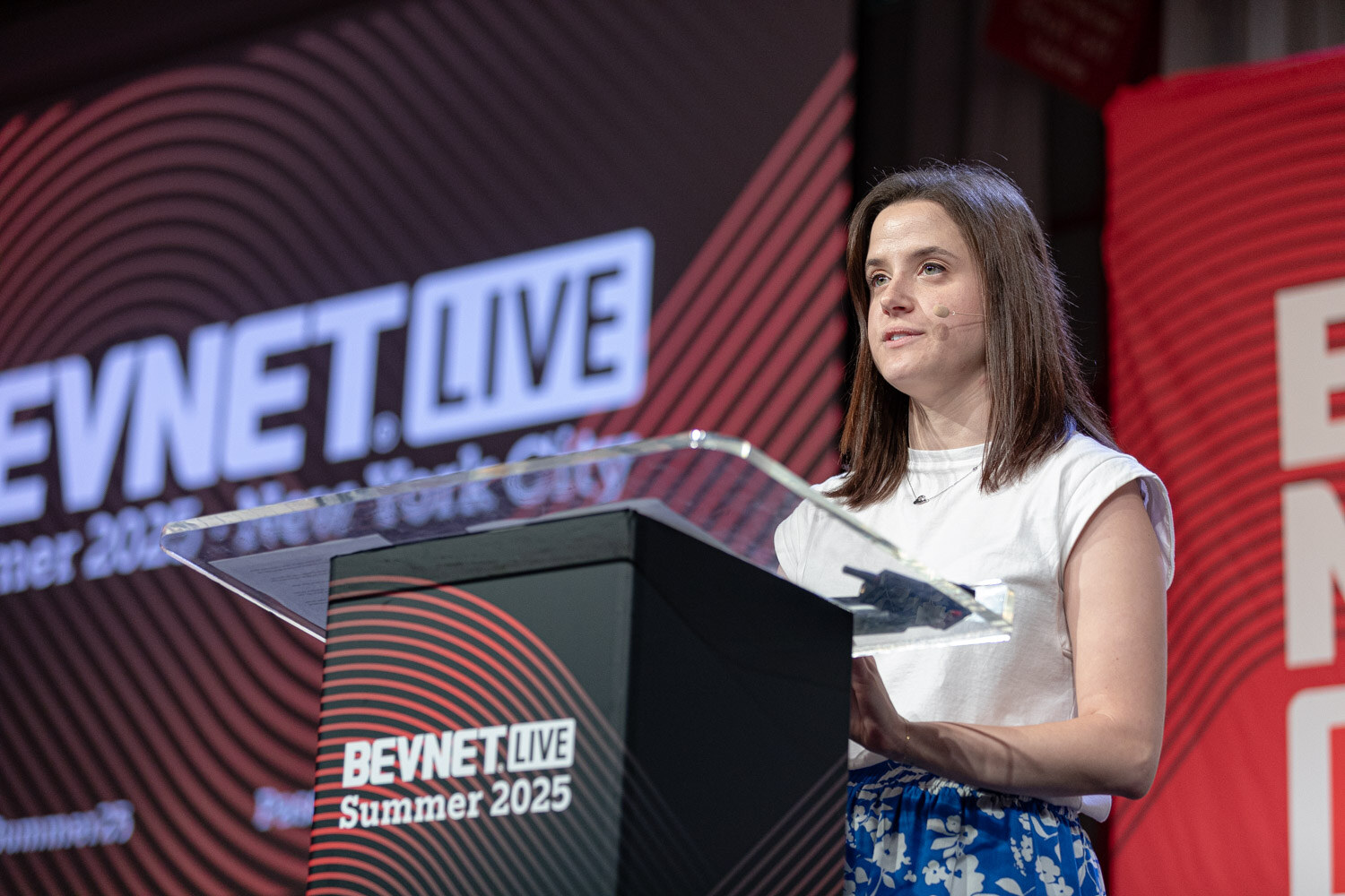 A woman in a white top and blue floral skirt speaks at a podium marked BevNET Live Summer 2025.