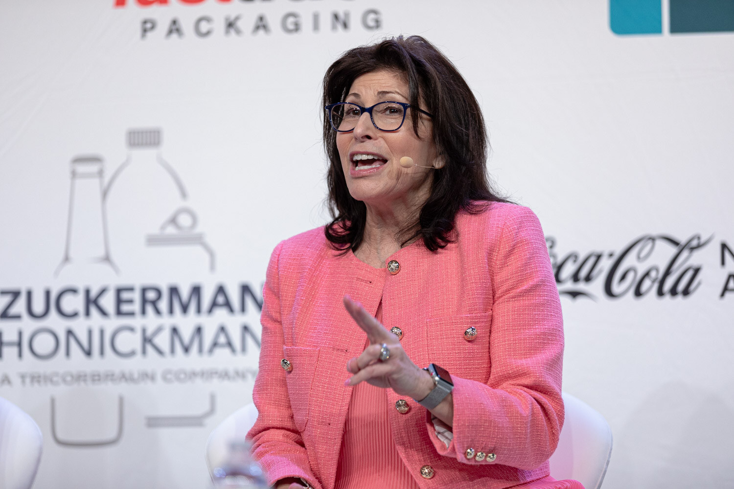 A woman in a coral jacket points one finger while seated in front of a white sponsor backdrop.