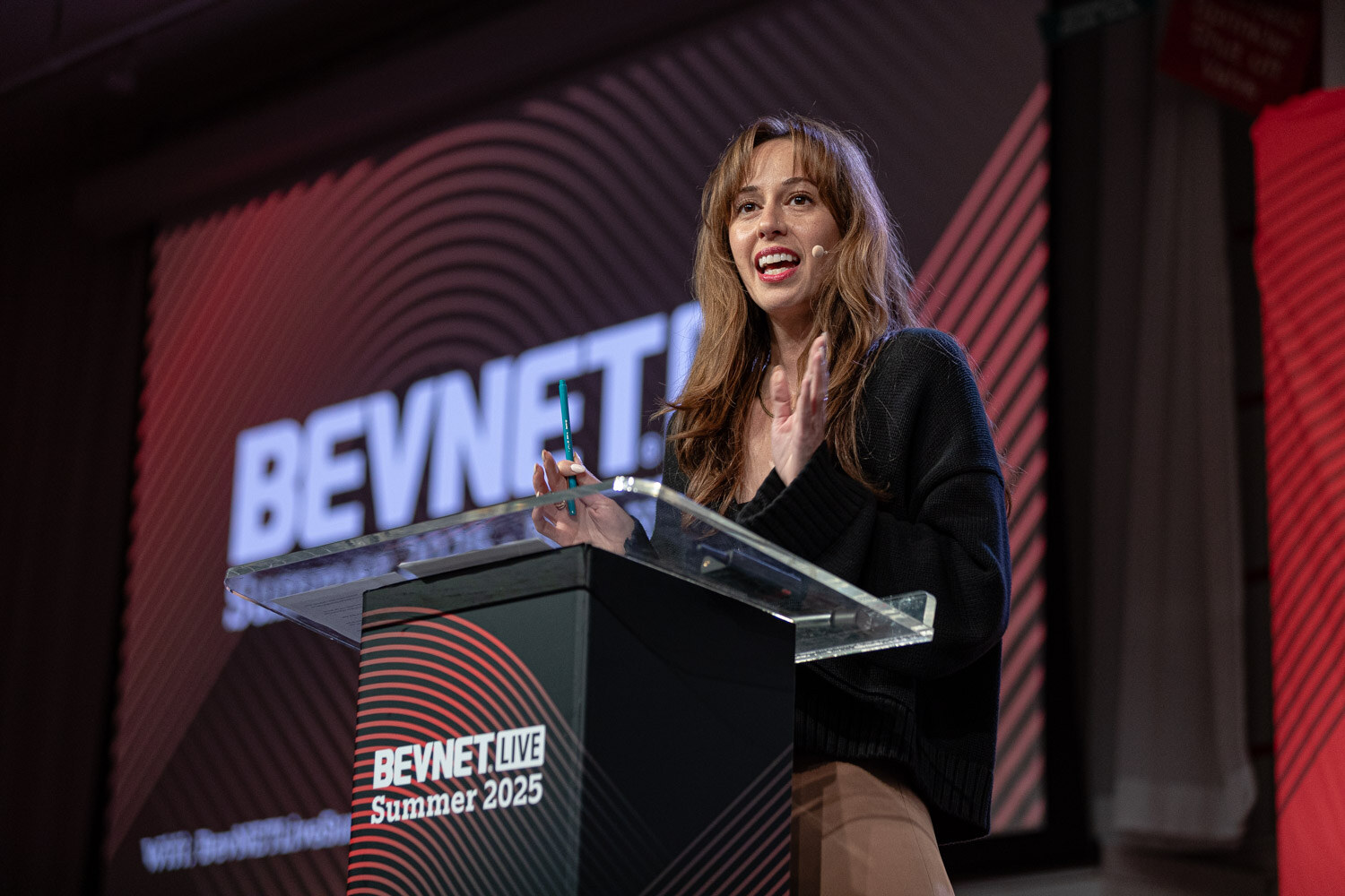 A woman with long brown hair in a black top speaks at a podium in front of a BevNET Live screen.