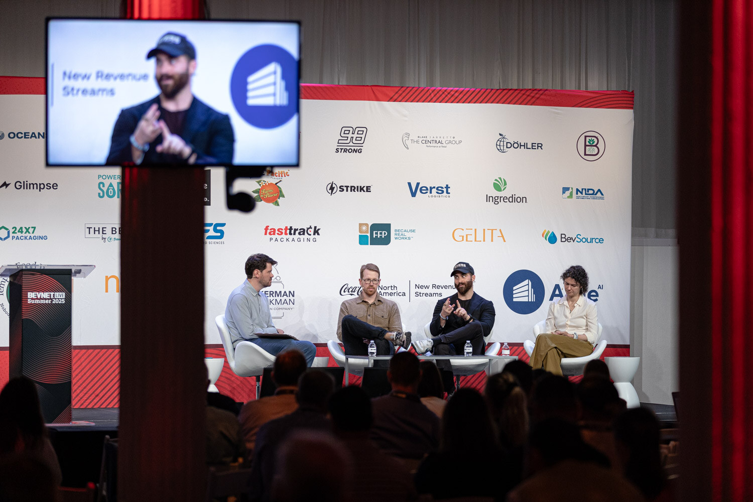 A wide shot of a four-person panel seated on stage, with a panelist shown on the side screen and an audience in the foreground.