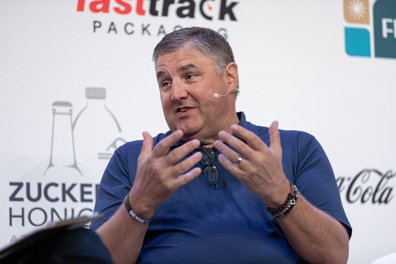 A man in a navy polo shirt gestures with both hands in front of a sponsor backdrop.