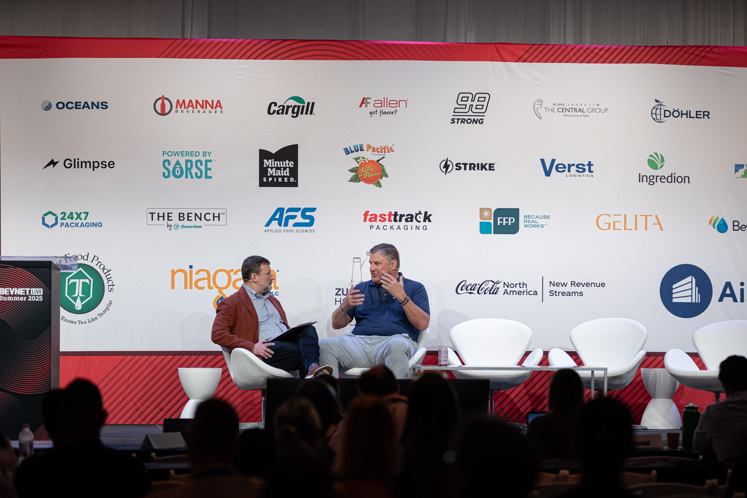 A wide shot of two men seated on a stage in front of a wall of sponsor logos, with audience silhouettes in front.