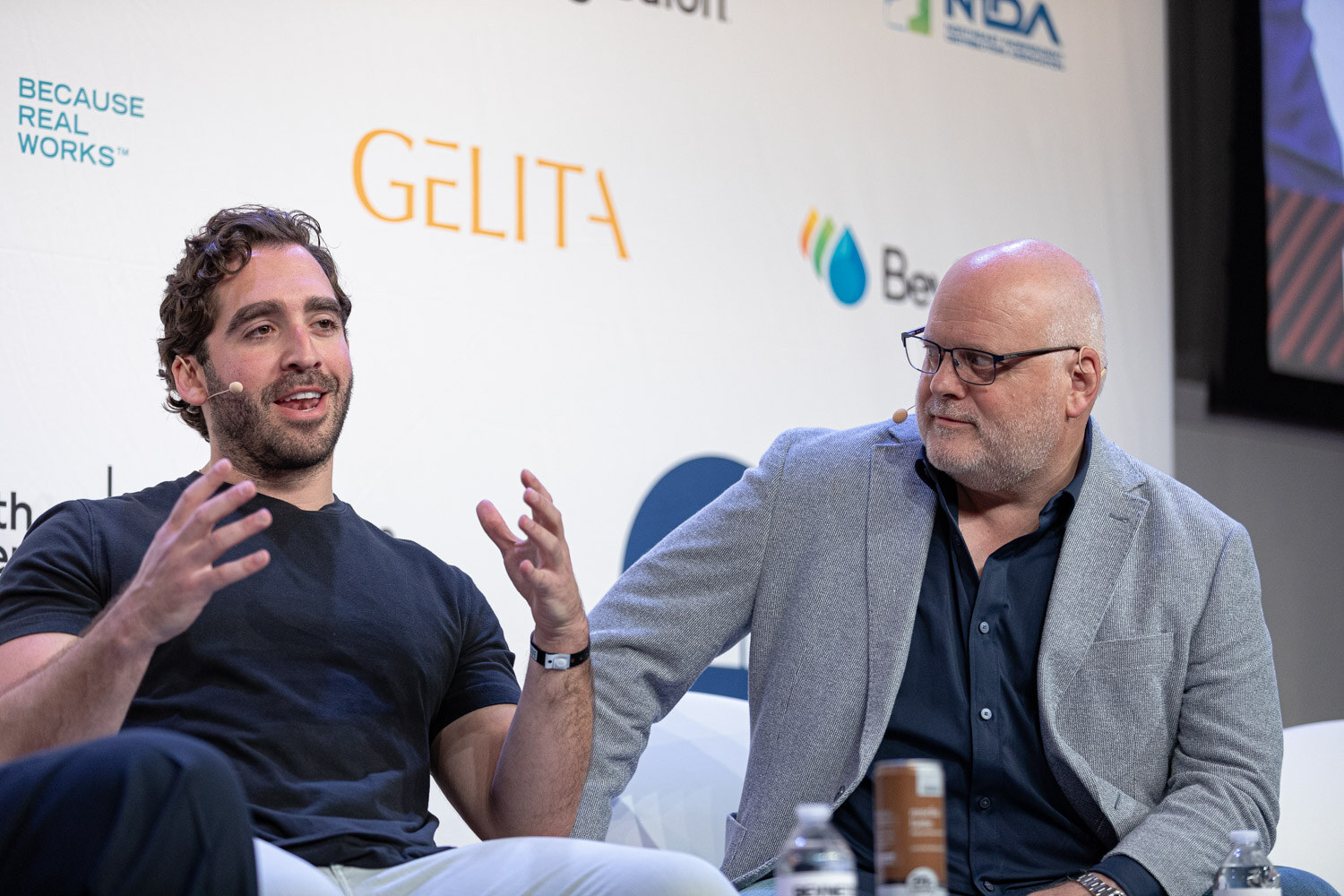 A dark-haired man in a black t-shirt gestures next to a bald man in a gray blazer, with sponsor logos behind them.