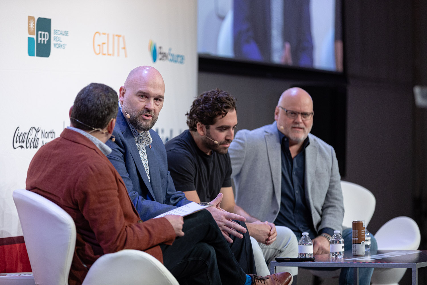 Four men sit in white chairs on stage, the moderator in a red-brown jacket facing the others.