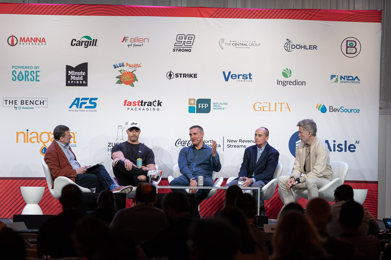 A wide shot of five people seated on a stage in front of a sponsor backdrop, with audience heads in the foreground.