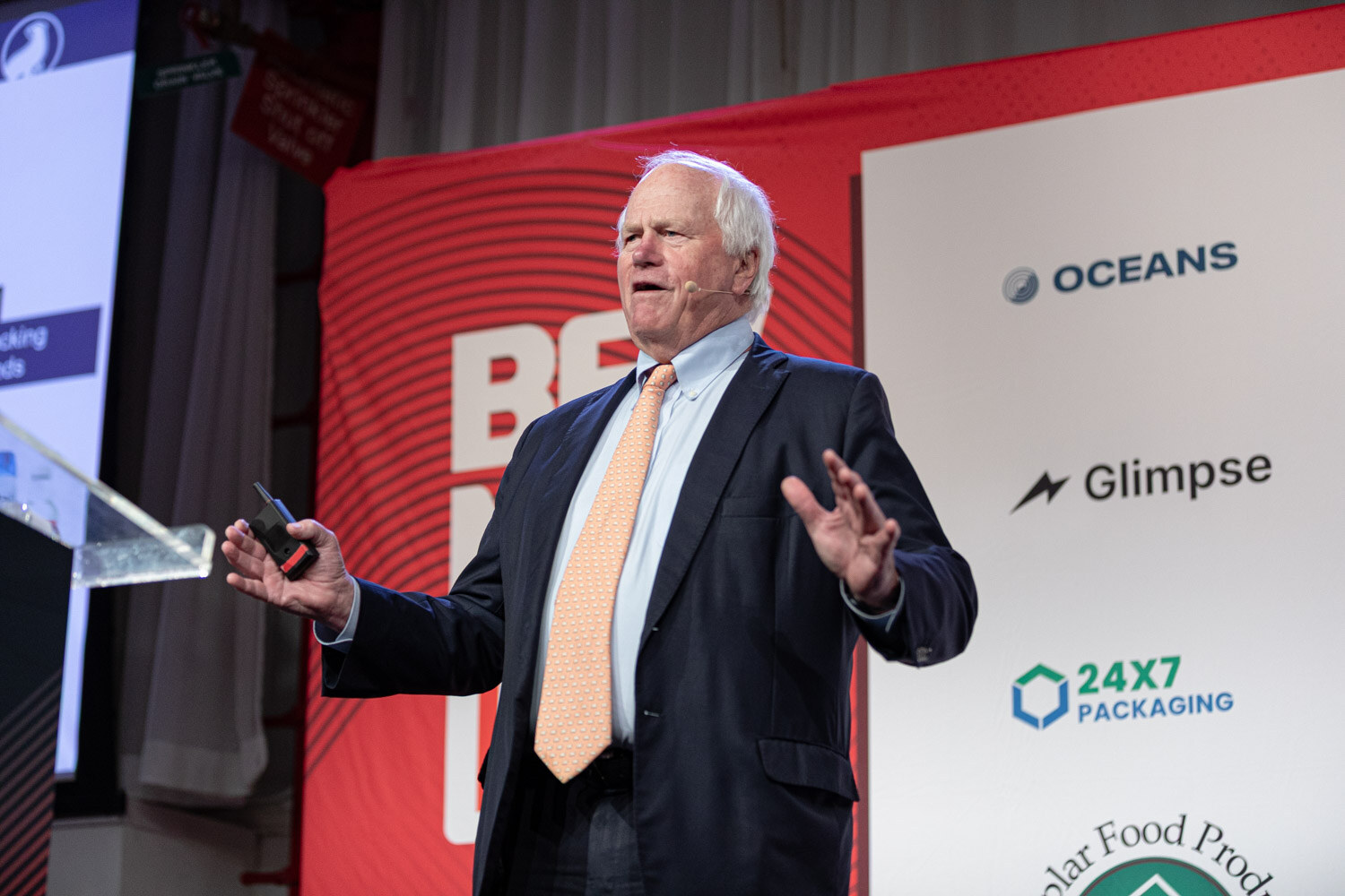 An older man in a dark suit and orange tie speaks with outstretched hands beside a podium and a red BevNET Live banner.