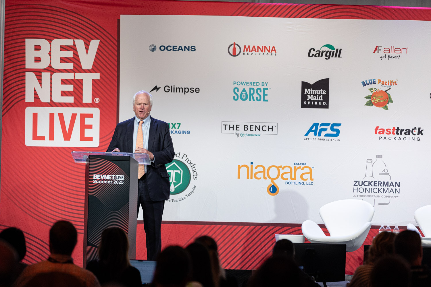 A wide shot of a man in a suit standing at a podium on a stage with a red BevNET Live banner and sponsor logos.