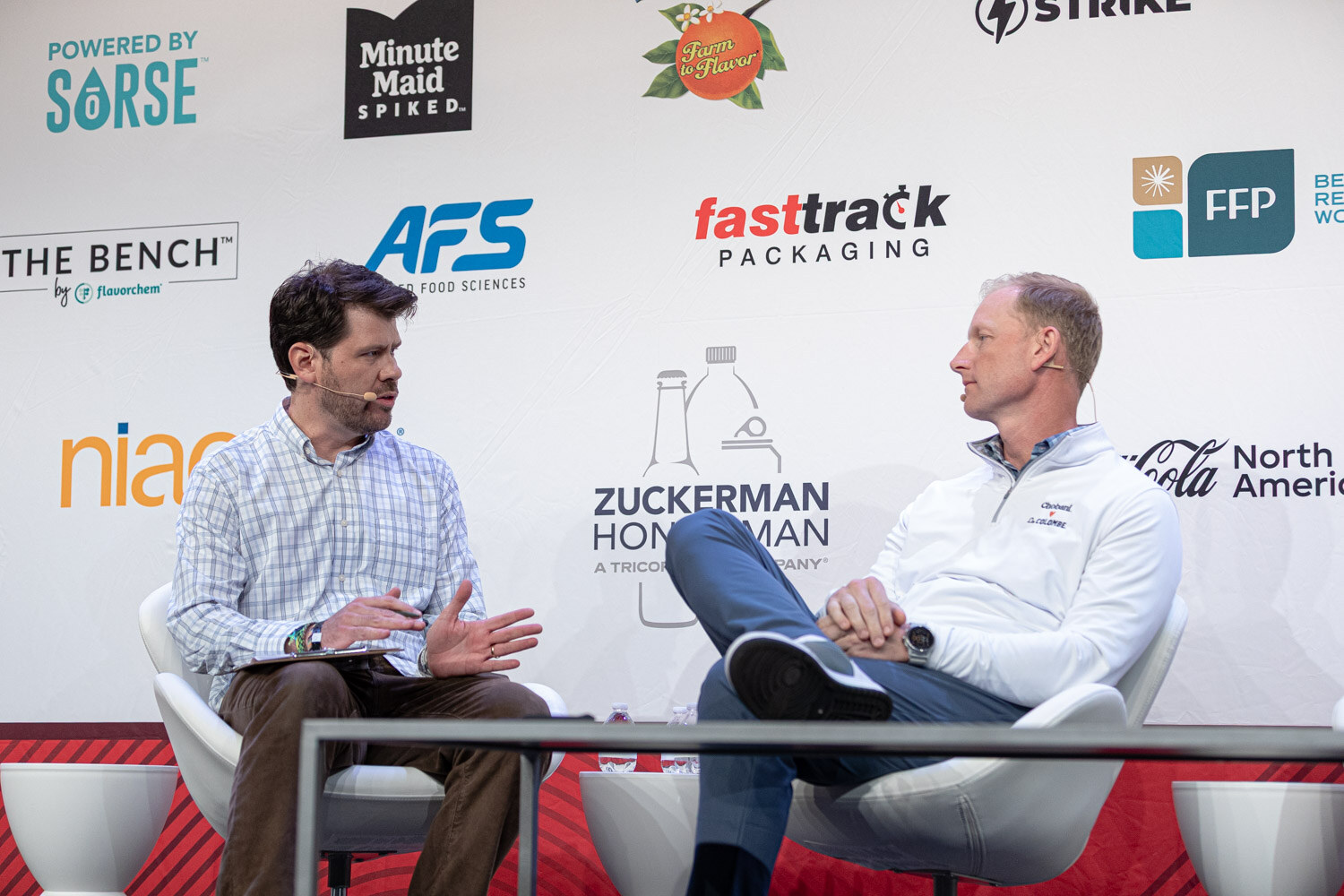 Two men sit on a stage in front of sponsor logos, one in a checked shirt and one in a white quarter-zip.