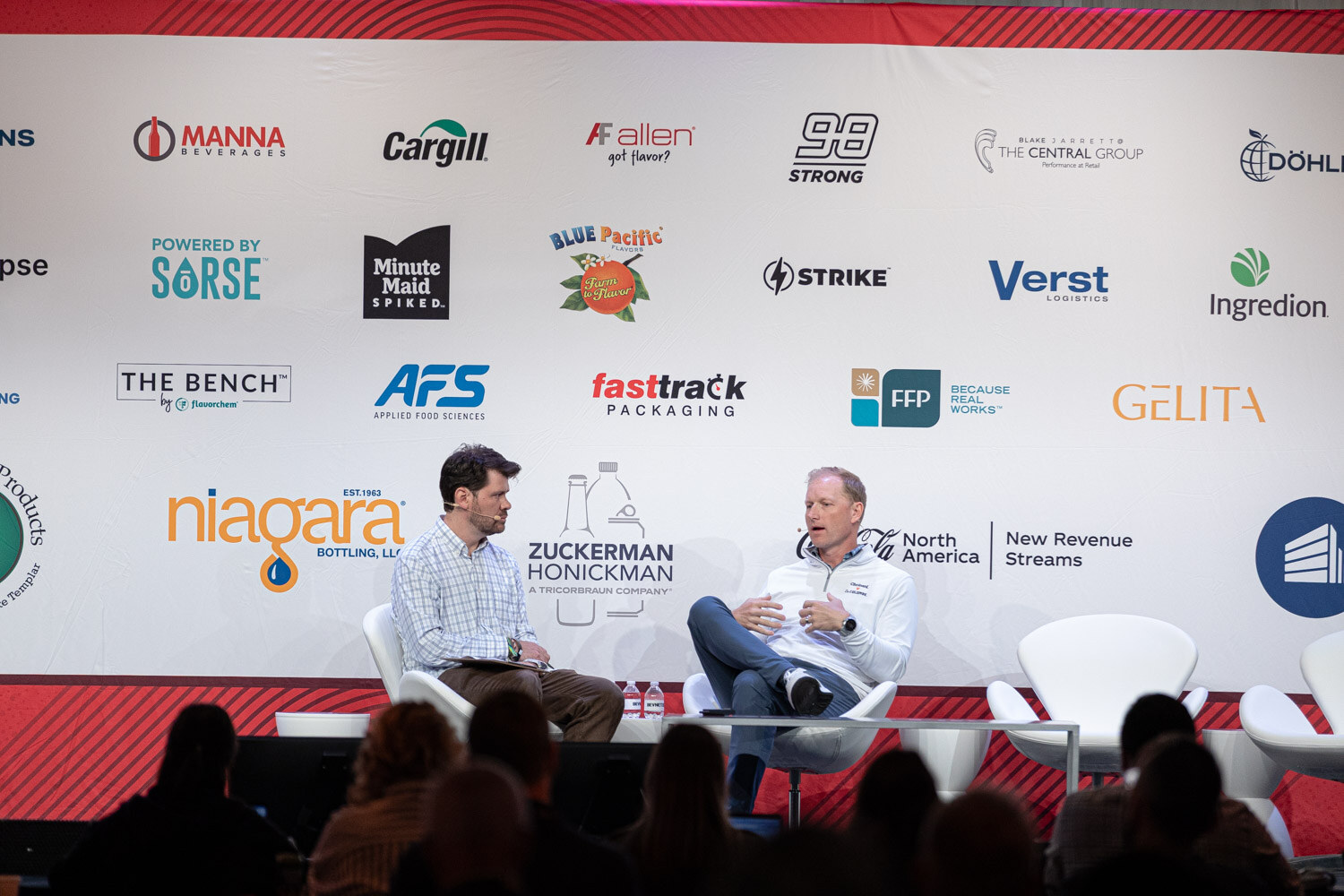 A wide shot of two men seated on stage in front of a sponsor wall, with audience silhouettes in front.