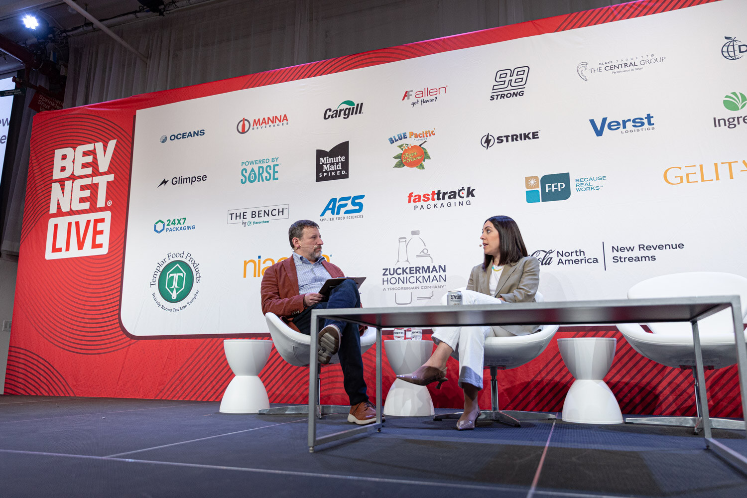 A wide shot of two people seated at a low table on a stage in front of a red BevNET Live banner and sponsor logos.