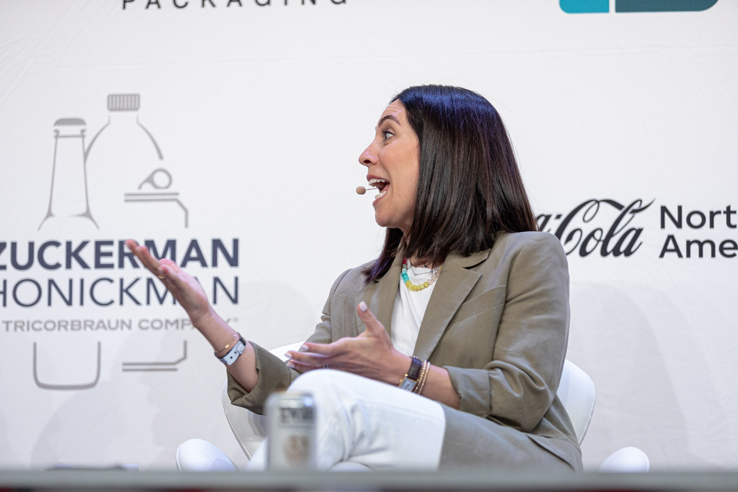 A dark-haired woman in an olive blazer gestures with both hands while seated in front of a Zuckerman Honickman sign.