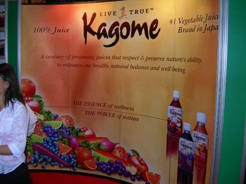 Booth wall reading Kagome, Live True, 100% juice, #1 Vegetable Juice Brand in Japan, with produce illustrations and bottles; a woman in a white top stands at the left edge.