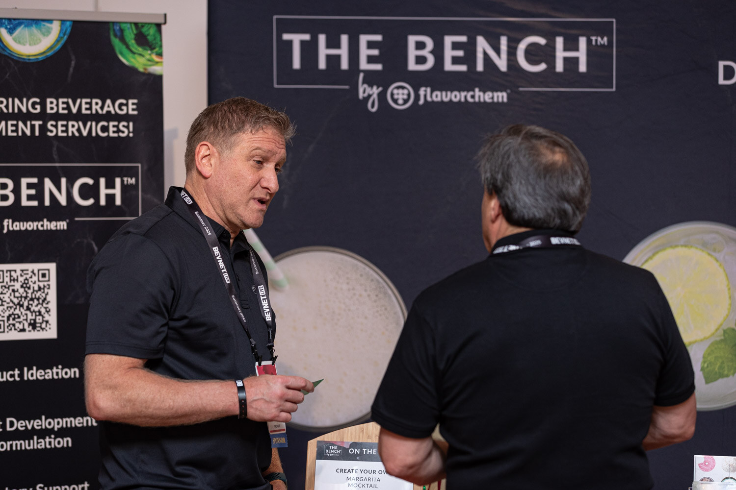 A man in a black polo and lanyard talks with another man in front of a The Bench by Flavorchem banner.