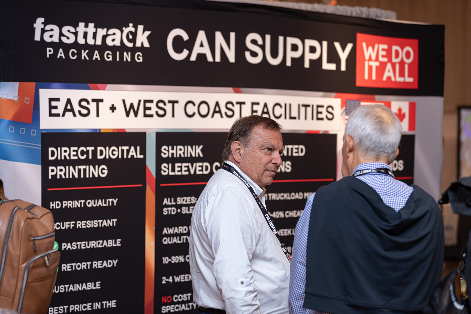 Two men with lanyards stand in front of a Fasttrack Packaging Can Supply banner.