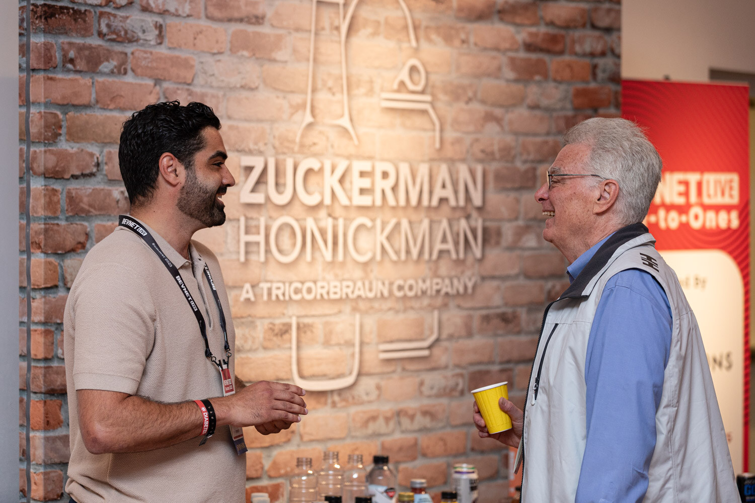 Two men with lanyards and yellow cups talk in front of a brick wall with a Zuckerman Honickman sign.