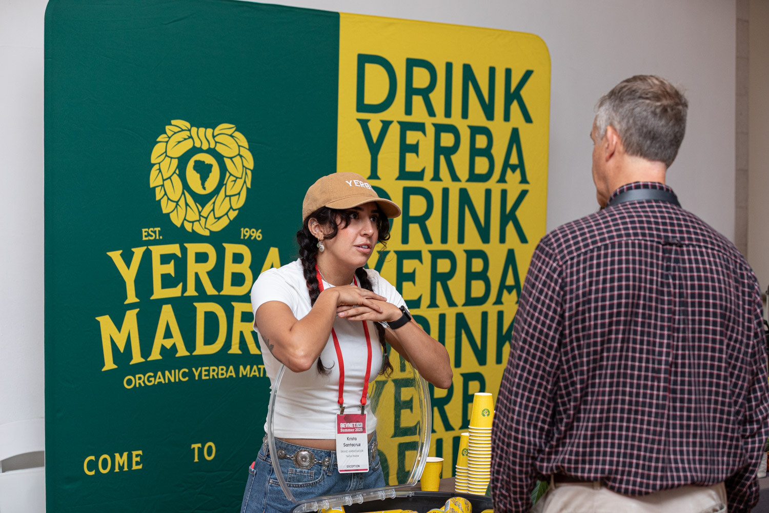 A woman in a cap talks with a man in a plaid shirt in front of a green and yellow Drink Yerba Madre banner.