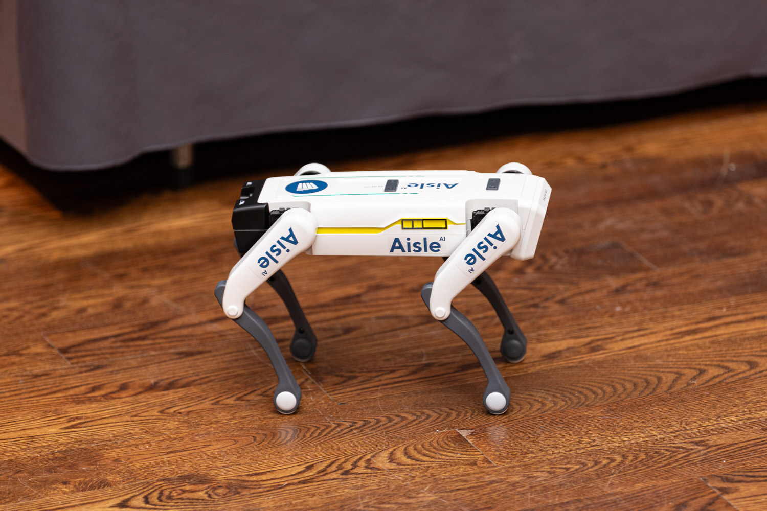 A white four-legged robot with Aisle printed on its legs stands on a wooden floor.