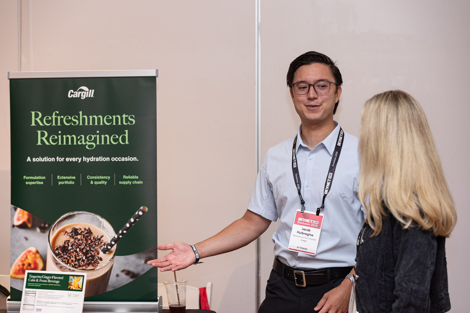 A man in a light blue shirt gestures toward a Cargill Refreshments Reimagined banner while a woman looks on.