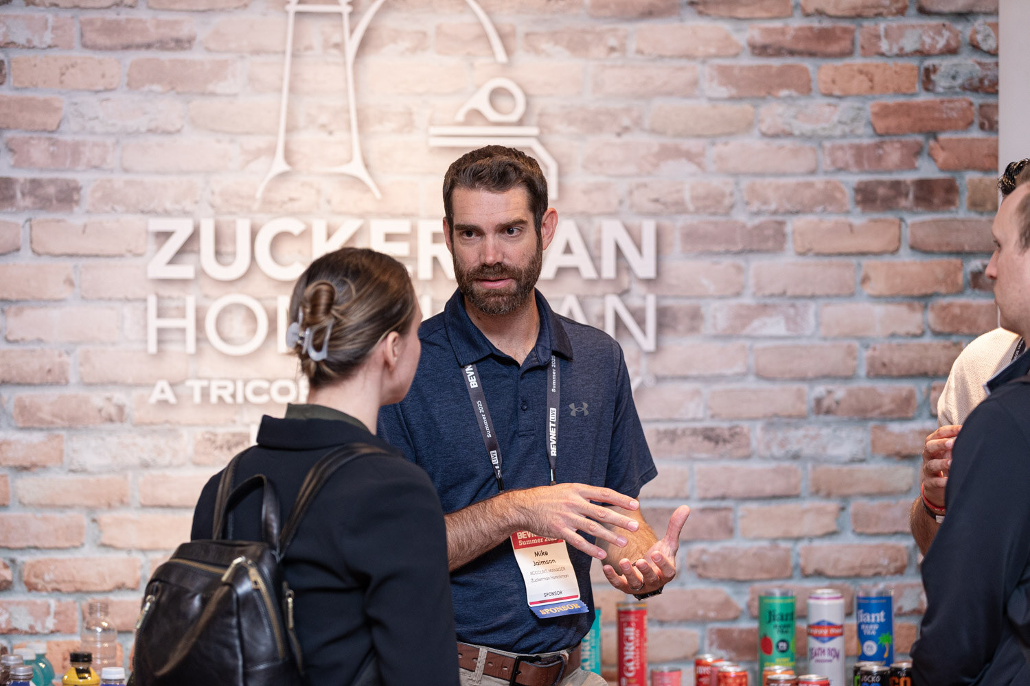A man in a navy polo gestures in front of a Zuckerman Honickman brick wall sign, with cans on a table.