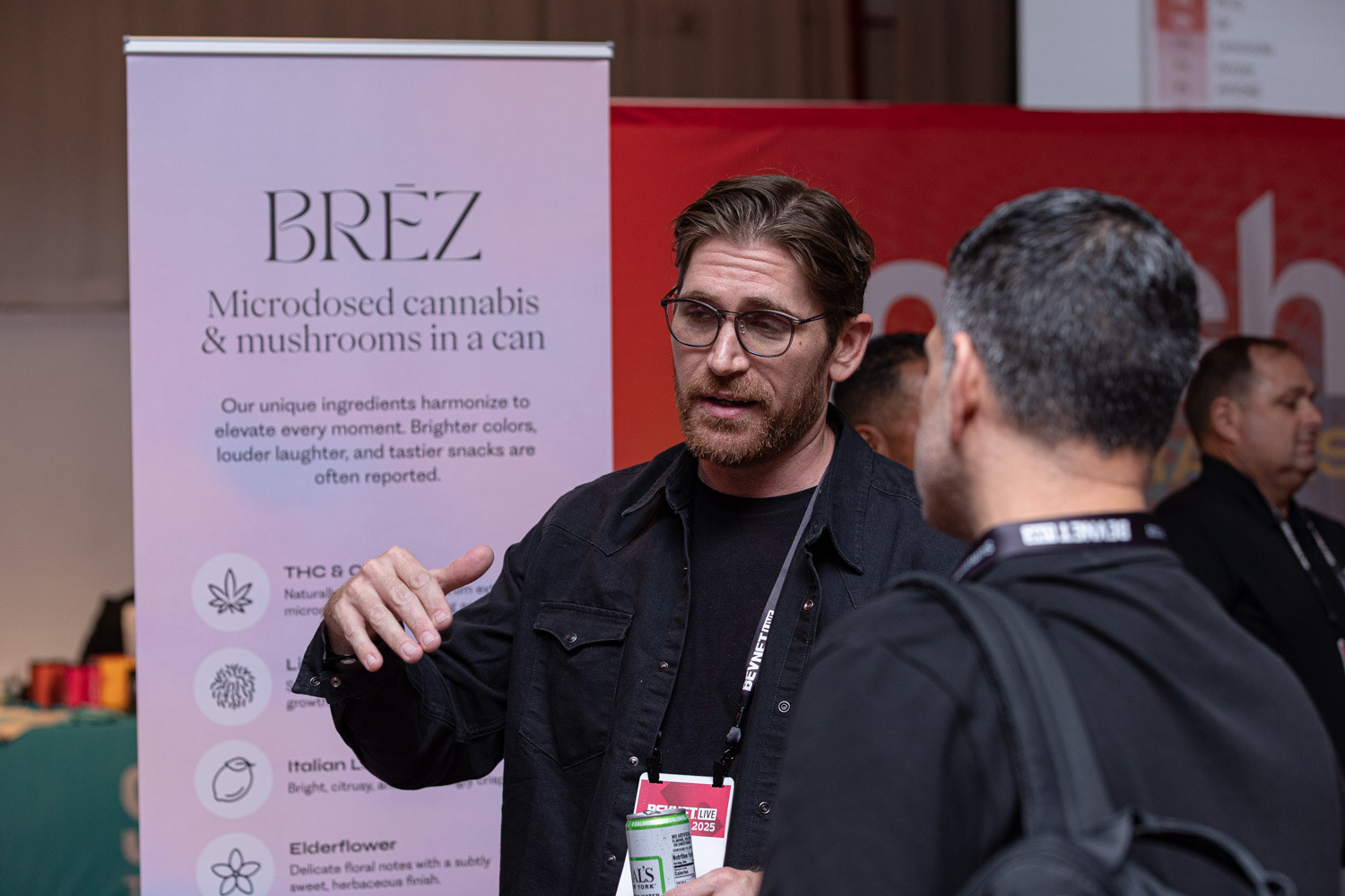 A bearded man in a dark jacket gestures in front of a Brez Microdosed cannabis and mushrooms in a can banner.