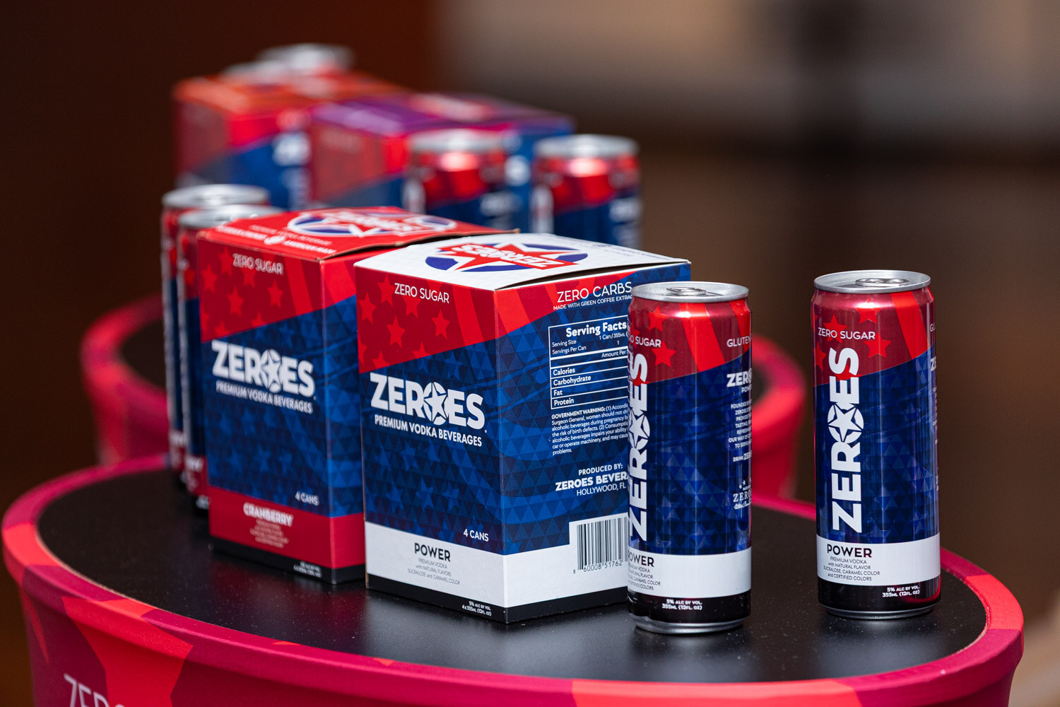Red, white and blue Zeroes cans and four-pack boxes on a round table.