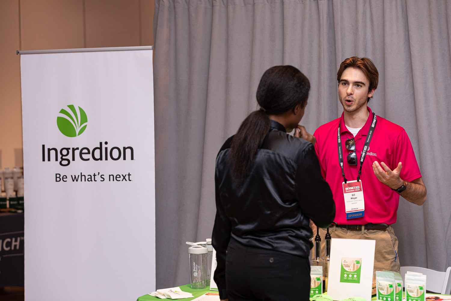 A man in a pink polo talks with a woman in black in front of an Ingredion Be what's next banner.