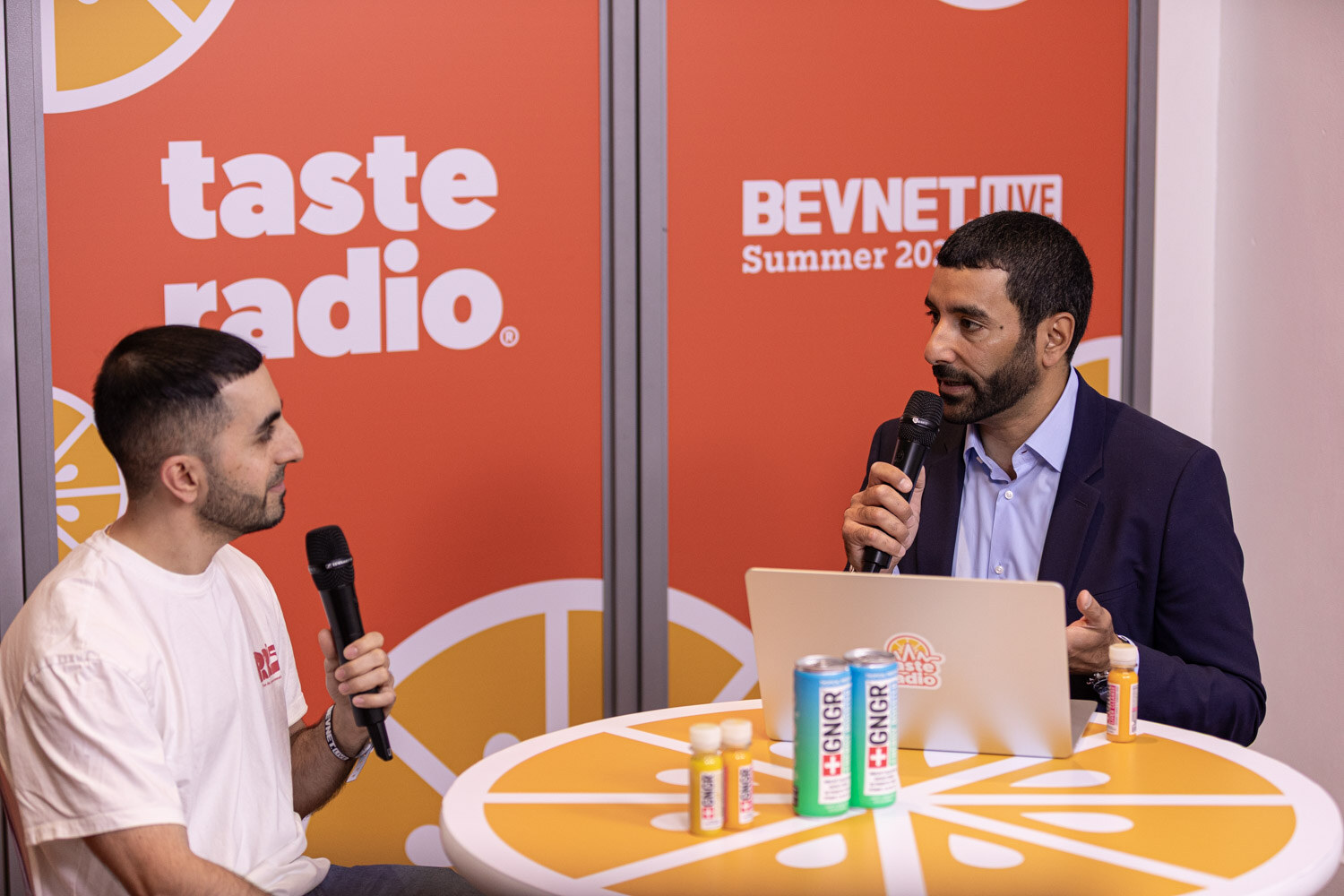 Two men with microphones sit at a round orange table in front of a Taste Radio and BevNET Live backdrop, with cans and shot bottles on the table.