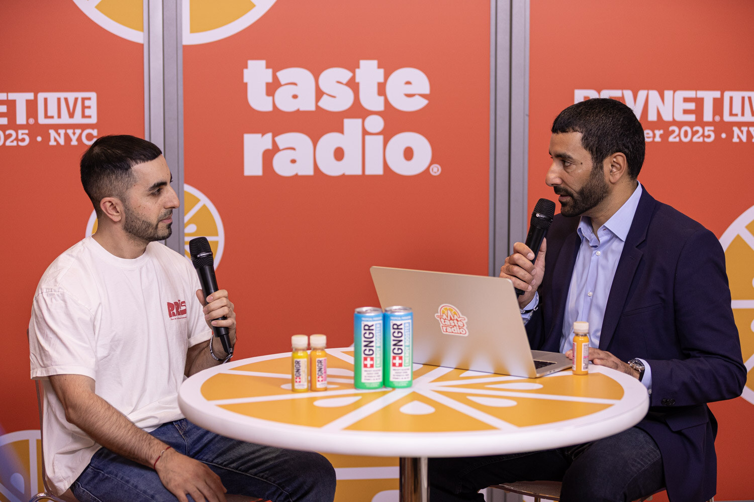 A man in a white t-shirt and a man in a navy blazer hold microphones at a table with GNGR cans and a laptop.