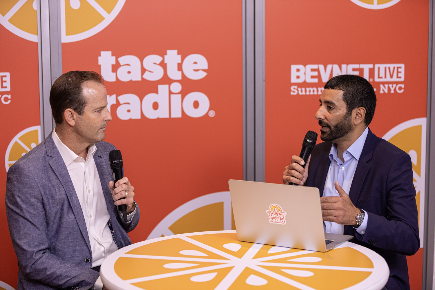 A man in a gray blazer and a man in a navy blazer hold microphones at a table with a laptop in front of an orange Taste Radio wall.