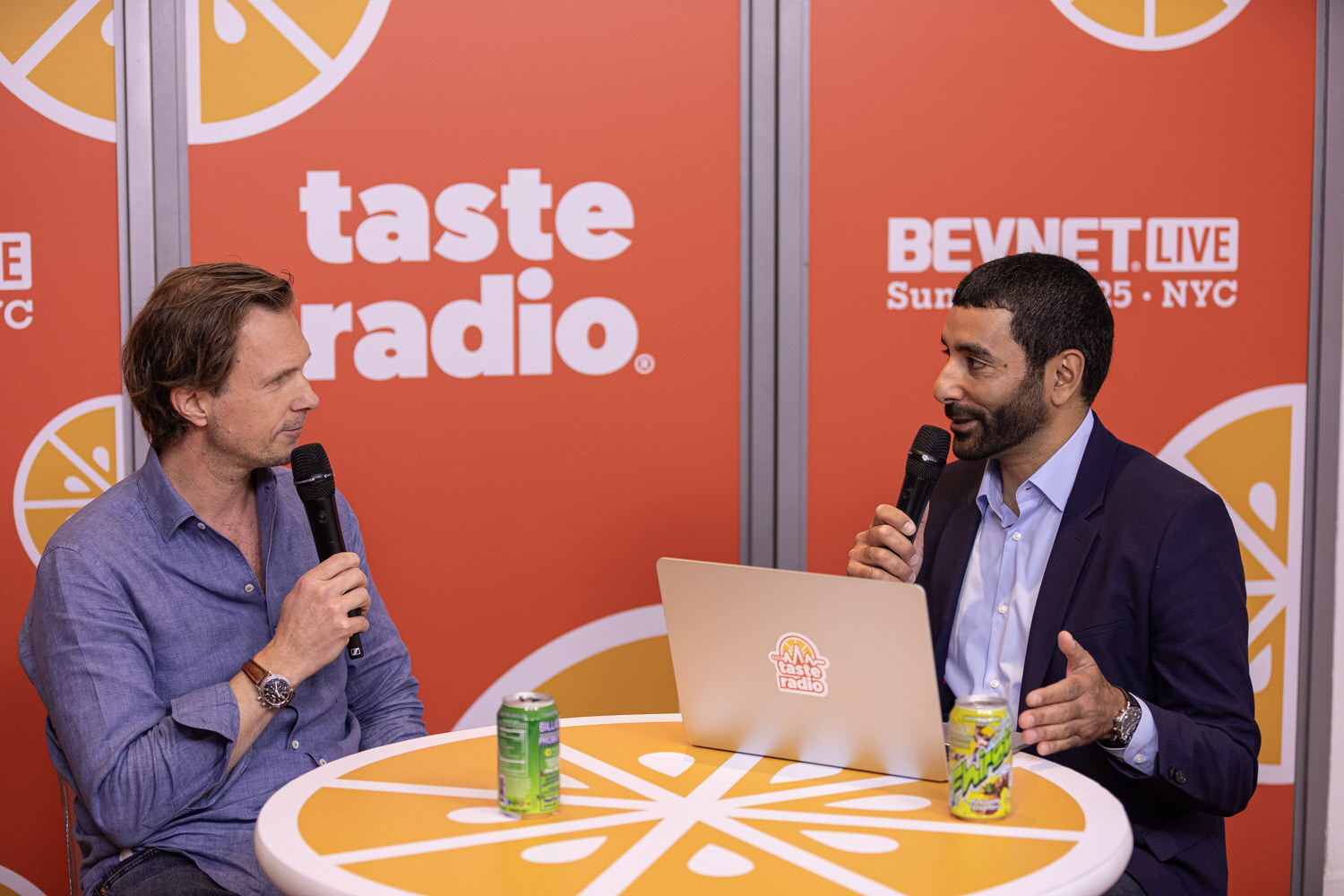 A man in a blue shirt and a man in a navy blazer hold microphones at a round table with two green cans and a laptop.
