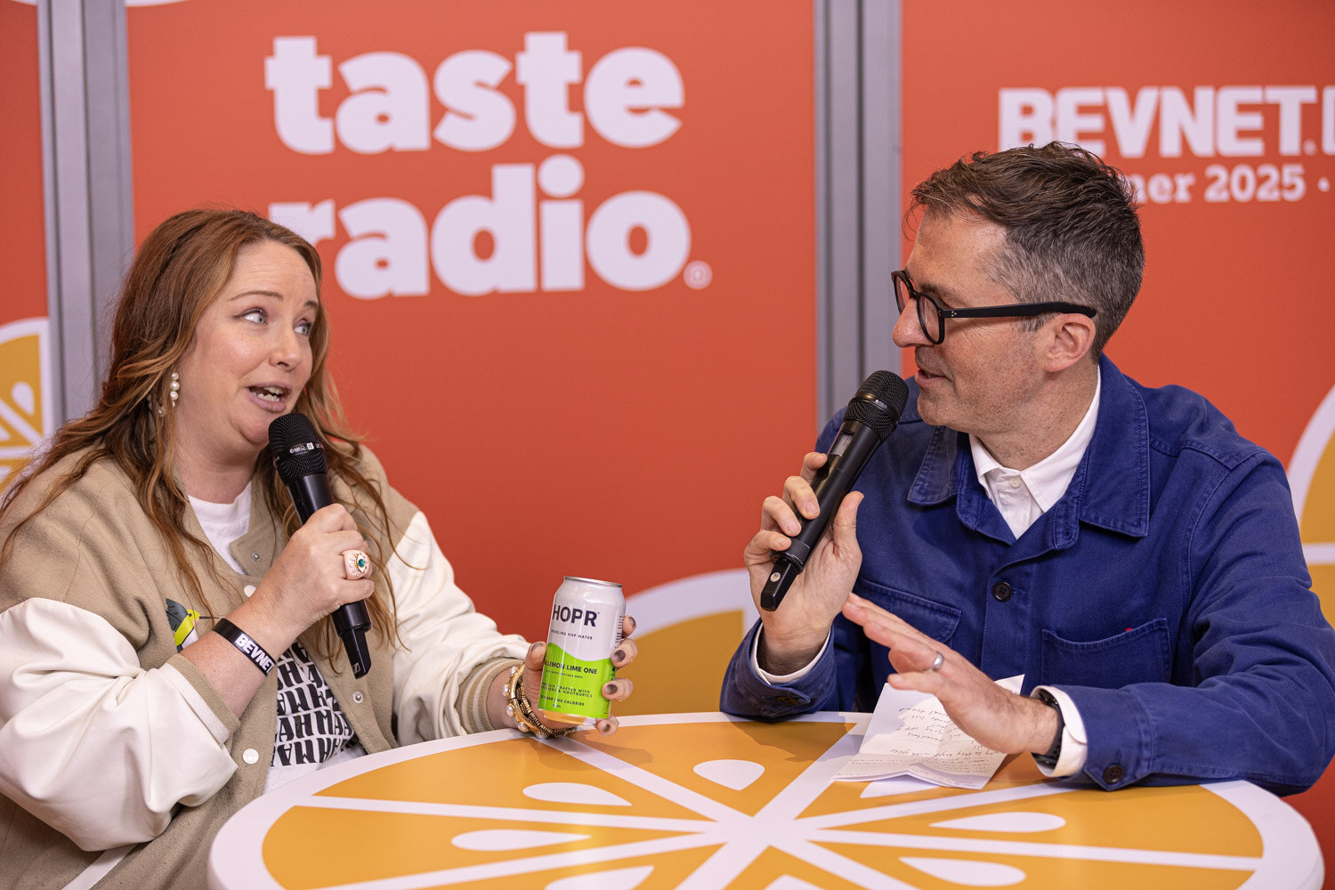 A woman in a beige and white jacket holds a green HOPR can and a microphone across from a man in a blue jacket.