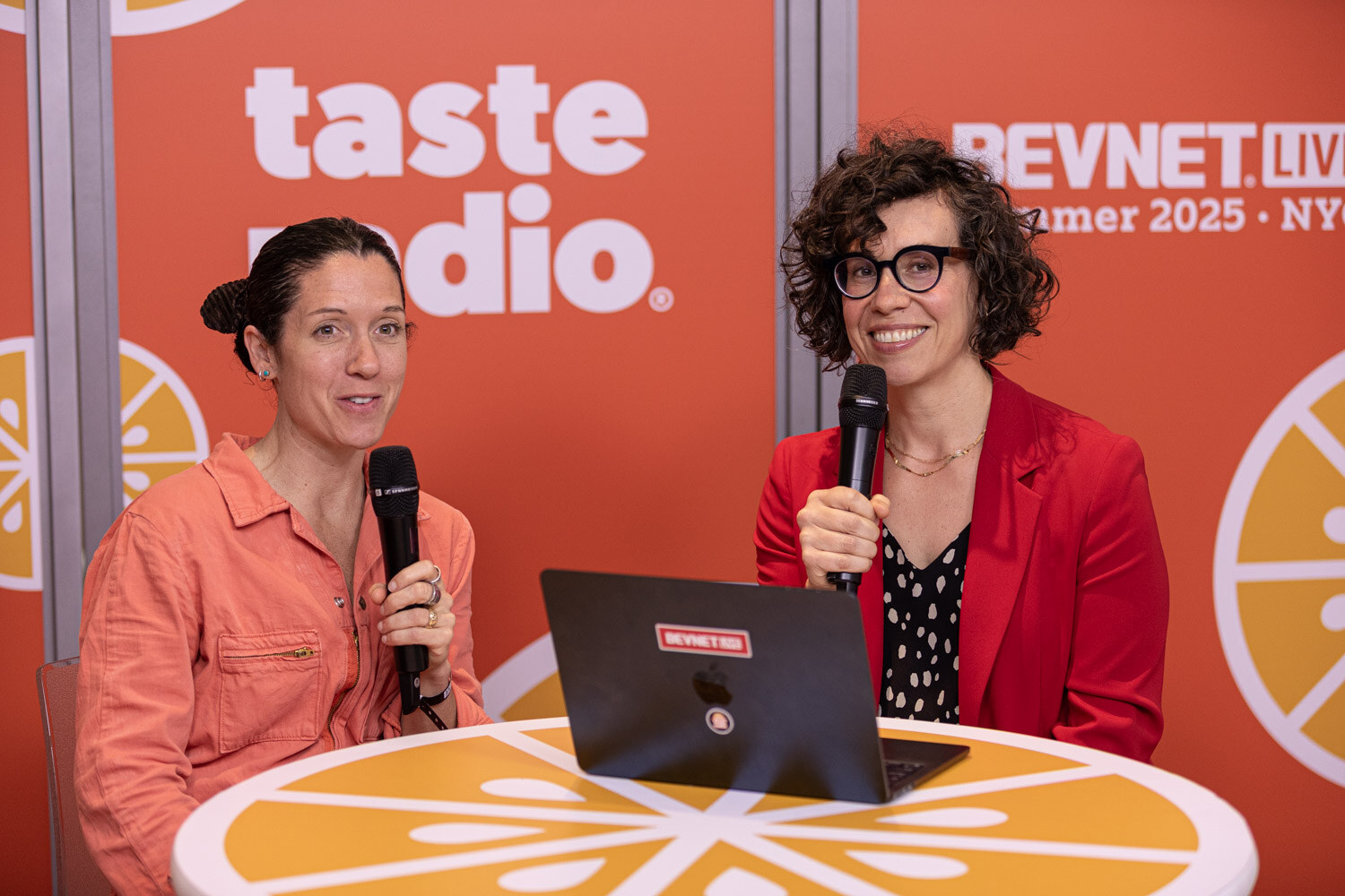 A woman in an orange jumpsuit and a woman in a red blazer hold microphones at a table with a laptop.