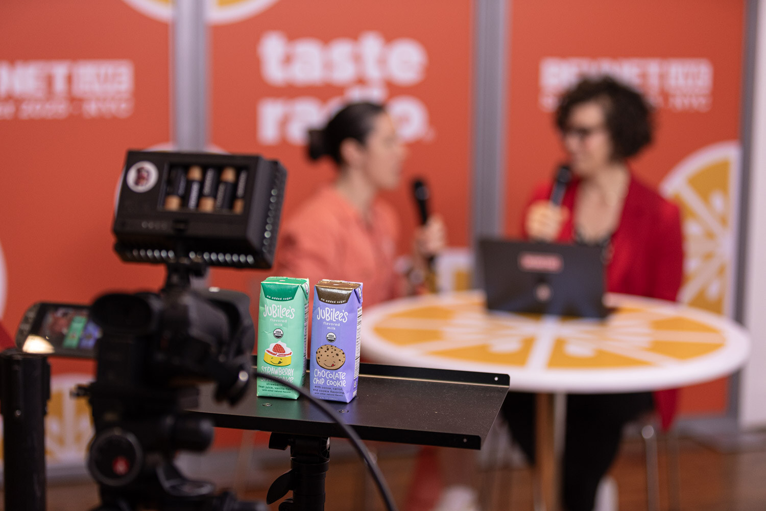 A video camera and two drink cartons on a stand in the foreground, with two people recording an interview at an orange table behind.